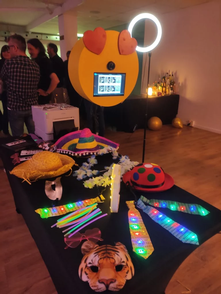 Fotomatón con cara de emoji y mesa negra llena de atrezo, corbatas, gorros, gafas, pelucas.
